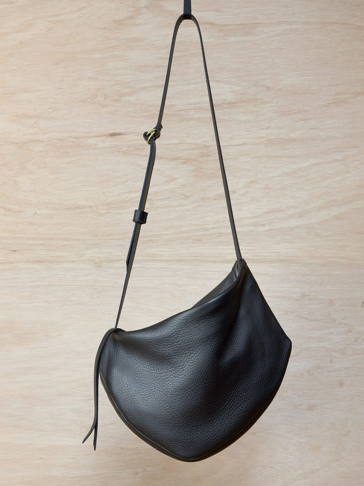 A Side Saddle black leather shoulder bag hanging on a wooden wall. Brand name: Kohl &amp; Co.