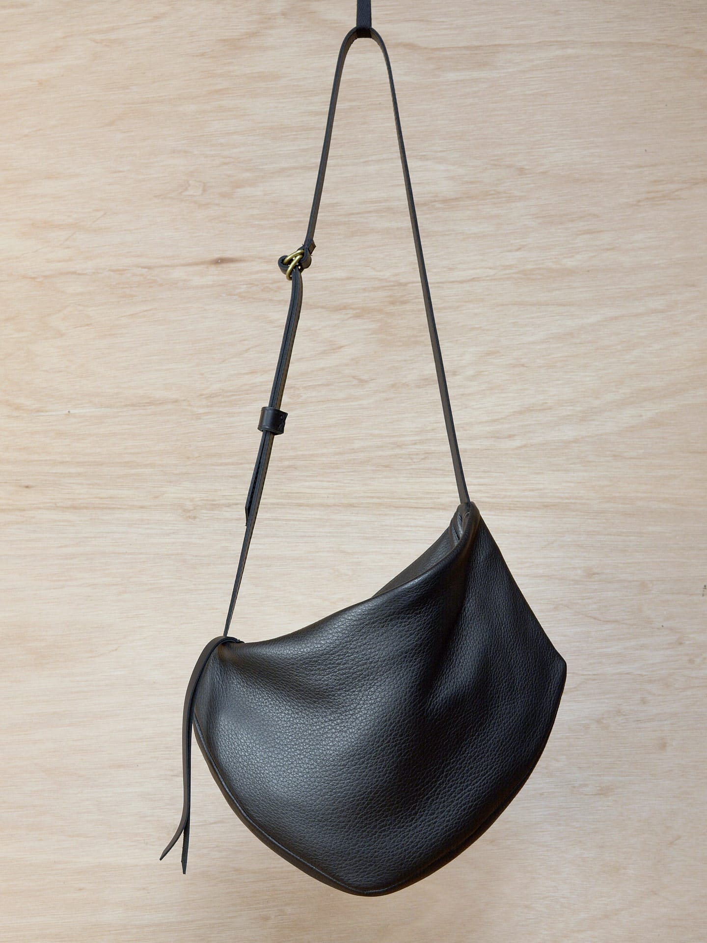 A Side Saddle black leather shoulder bag hanging on a wooden wall. Brand name: Kohl & Co.