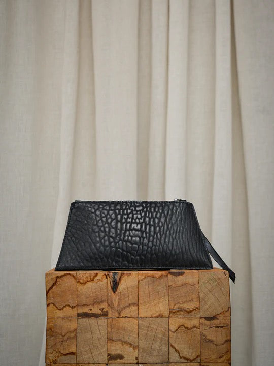 A Maya Carry – Black clutch from Kohl & Co sitting on top of a wooden block.