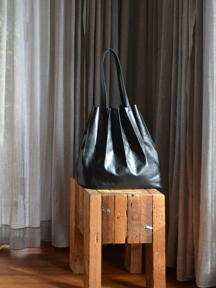A black leather Pleats bag sitting on a wooden stool. (Brand: Kohl & Co)