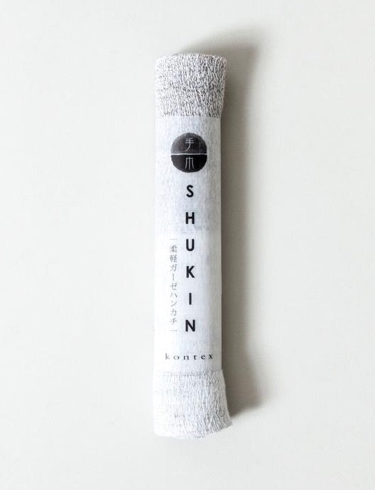 A folded white towel with Japanese text and the word &quot;Shukin Towel - Two Tone Grey ⋄ Pink&quot; on its packaging label, made in Imabari, against a light background by Kontex.