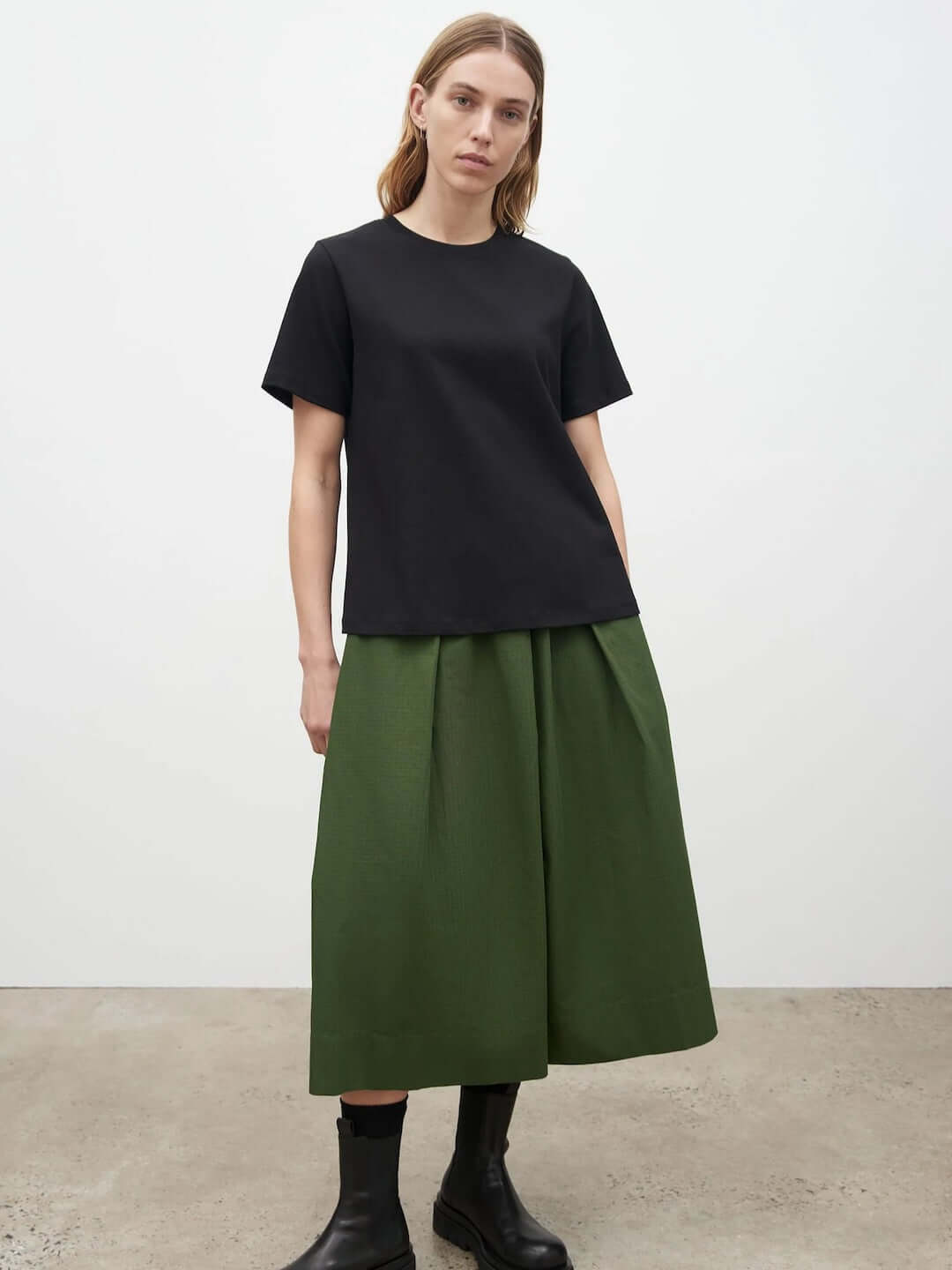 The model is wearing a A-Line Tee – Black by Kowtow and green pleated skirt.