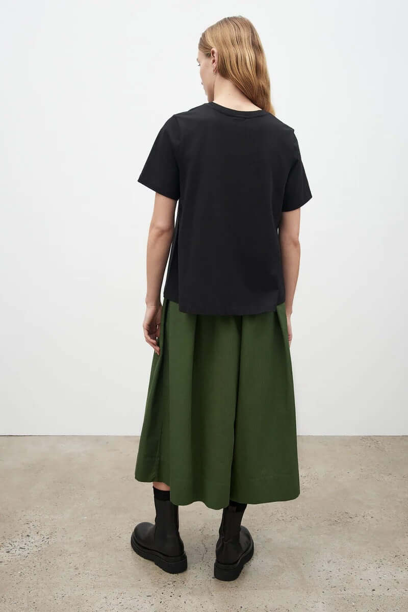 The back view of a woman wearing a &quot;A-Line Tee – Black&quot; from Kowtow and green pleated skirt.