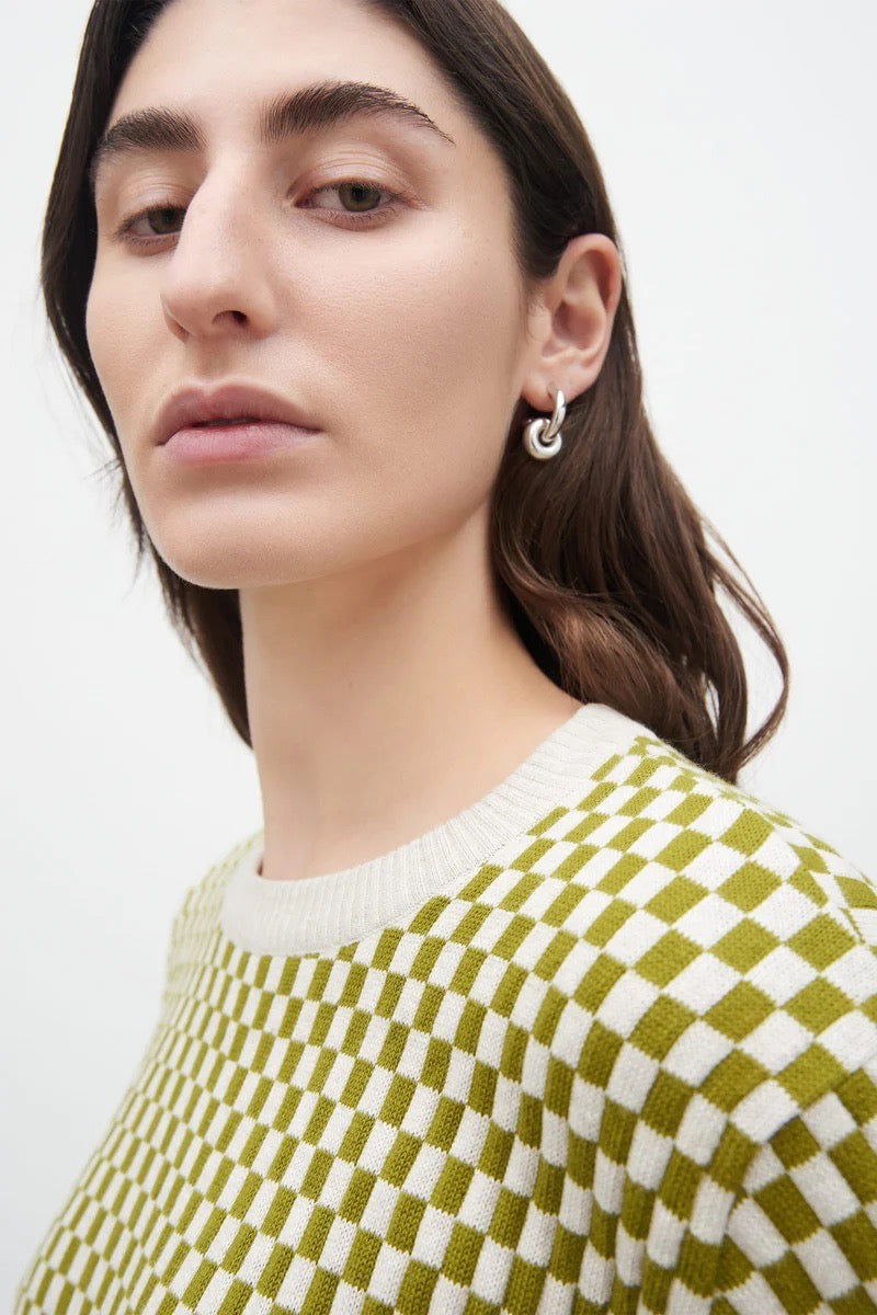 A woman wearing a Kowtow Checkerboard Knit Crew – Lawn Check sweater and earrings.