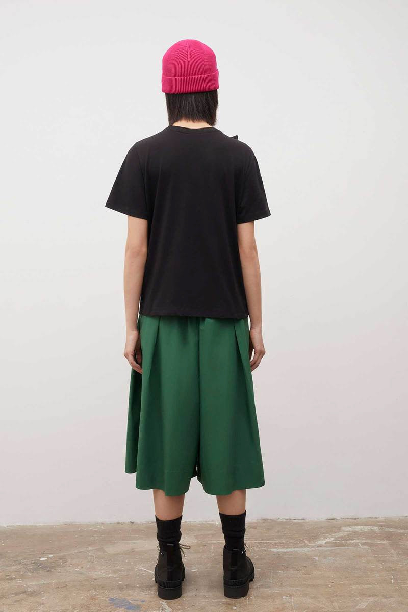 A woman wearing a Kowtow Knotted Tee - Black and green pleated skirt.