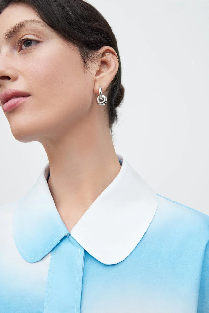 A woman wearing a blue Play Top - Cloud shirt and Kowtow white earrings.