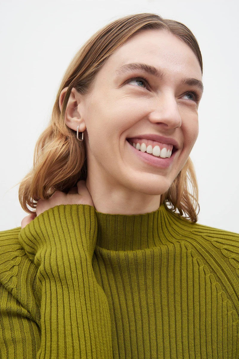 A woman wearing a Kowtow green Row Top - Lawn sweater and hoop earrings.