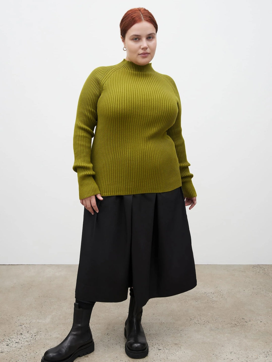 A woman wearing a green Row Top - Lawn turtleneck sweater and black skirt. (Brand Name: Kowtow)