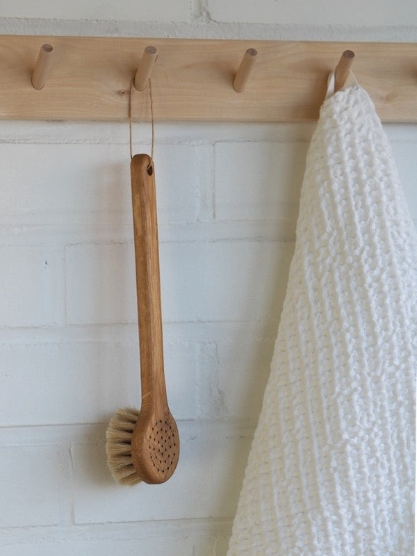A Body Brush – Round Head from Iris Hantverk hangs on a hook next to a white towel.
