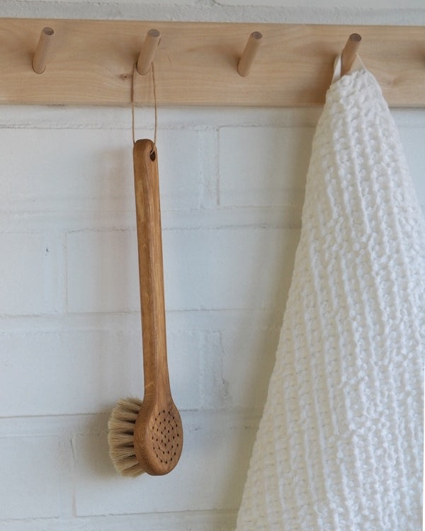 A Body Brush – Round Head from Iris Hantverk hangs on a hook next to a white towel.