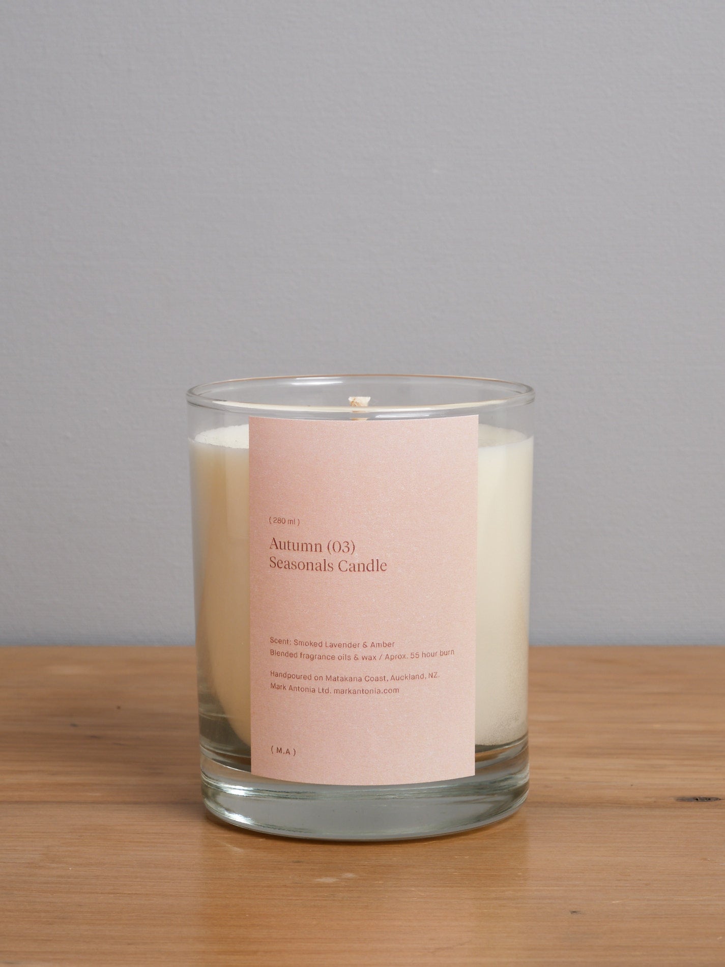 A Mark Antonia Seasonal Candle (2.0) – Autumn with a pink label sitting on a wooden table.