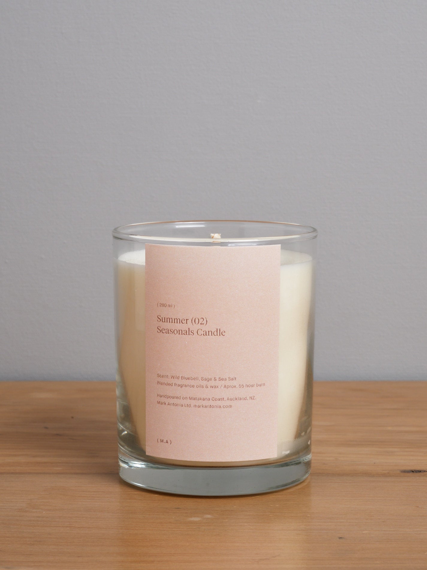 A Seasonal Candle (2.0) – Summer by Mark Antonia with a pink label on it sitting on a wooden table.