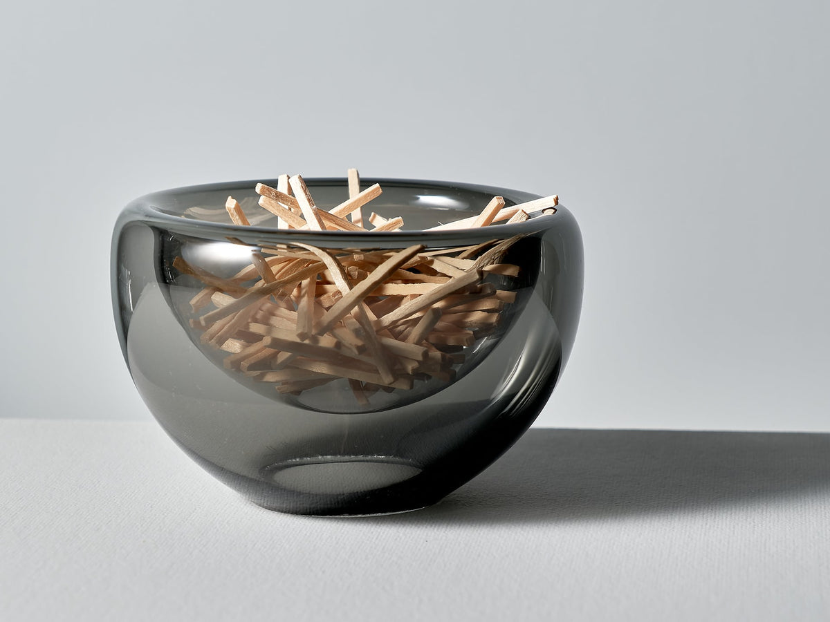 A Mini Fulvio Glass Bowl - Grey filled with match sticks on a white surface.