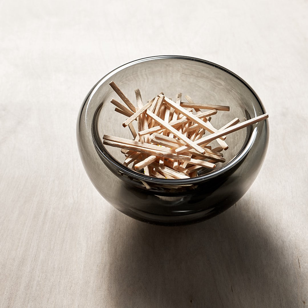 A Mini Fulvio Glass Bowl – Grey filled with wooden matchsticks on a wooden table. (brand: Matthew Hall)