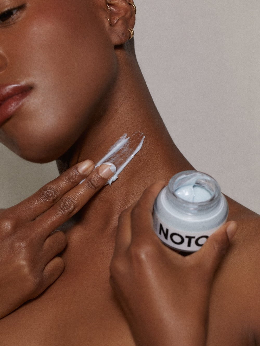 A woman is holding a jar of NOTO Moisture Riser Cream on her face.