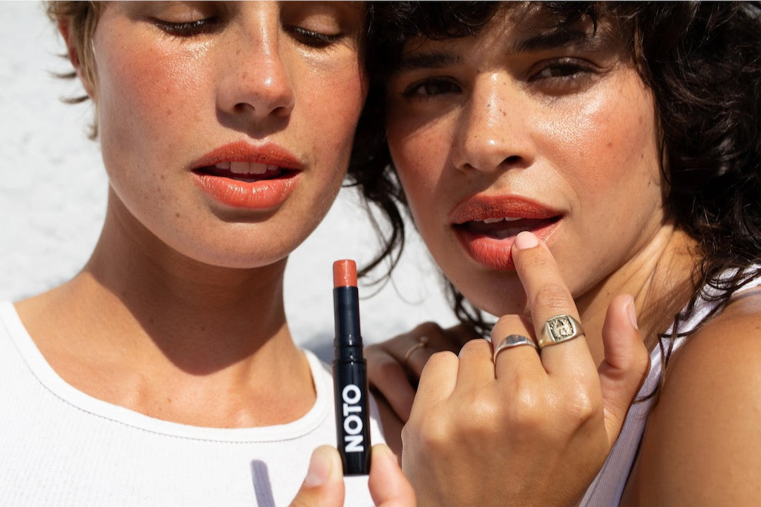 Two women are holding an Ono Ono – Multi-Bene Stain Stick // Lips + Cheeks by NOTO in their hands.