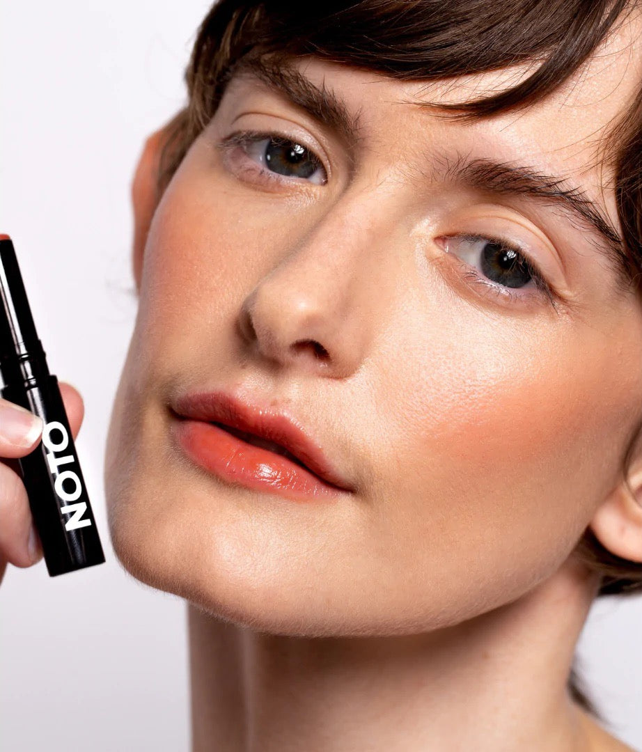 A woman is putting the NOTO Ono Ono – Multi-Bene Stain Stick // Lips + Cheeks on her lips.