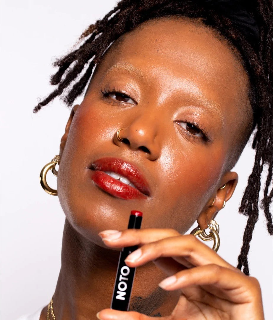 A woman with dreadlocks holding a NOTO Oscillate – Multi-Bene Stain Stick // Lips + Cheeks.