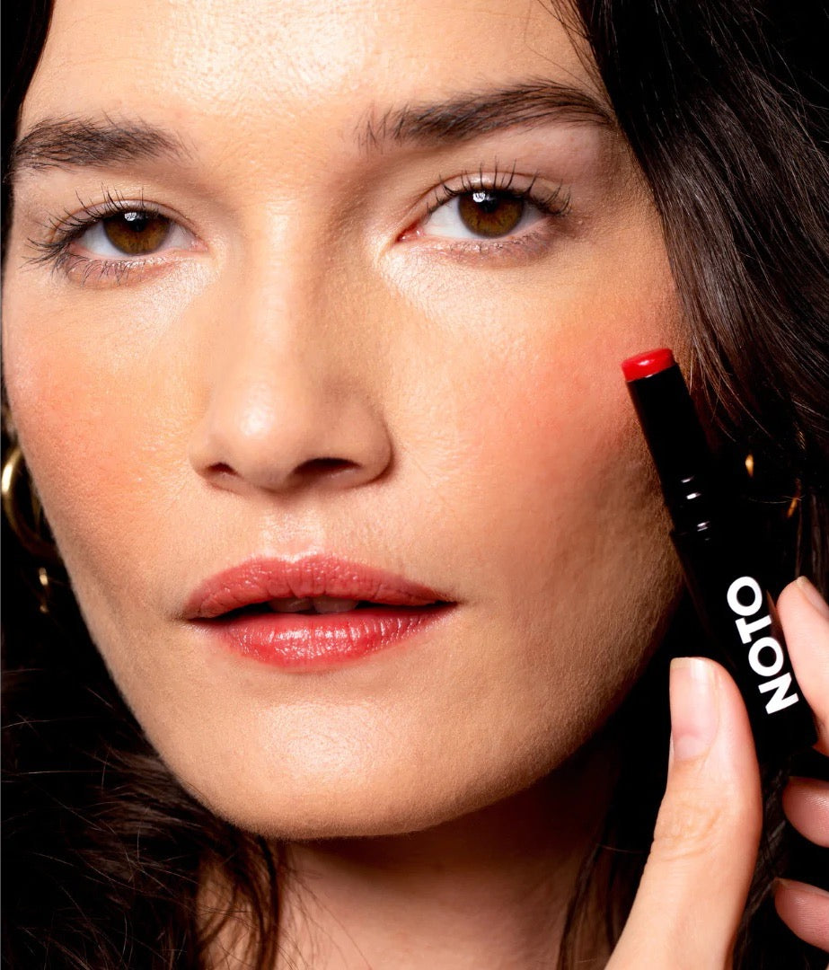 A woman is holding an Oscillate – Multi-Bene Stain Stick // Lips + Cheeks by NOTO in her hand.
