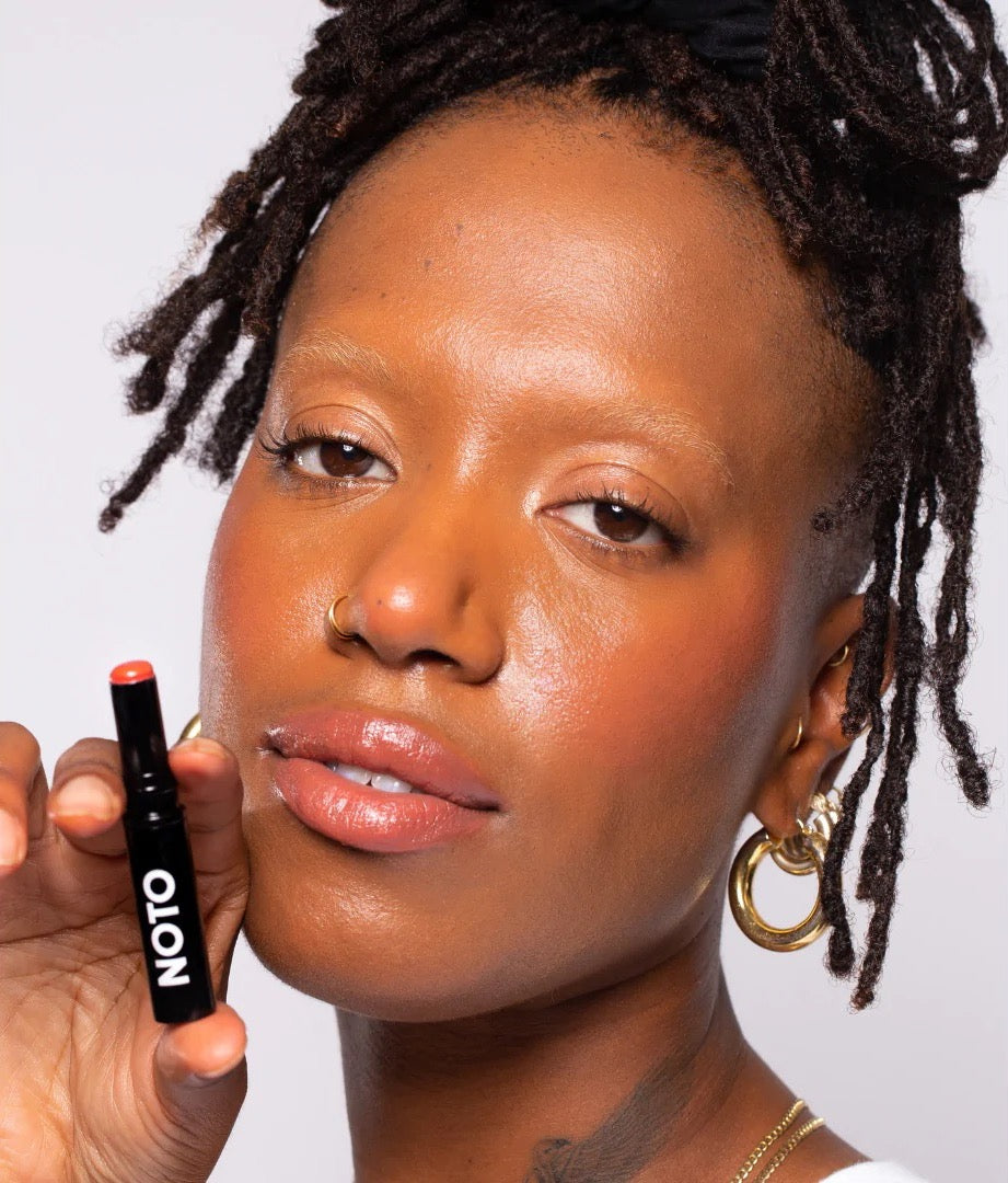 A woman with dreadlocks holding a tube of NOTO Touch – Multi-Bene Stain Stick // Lips + Cheeks.