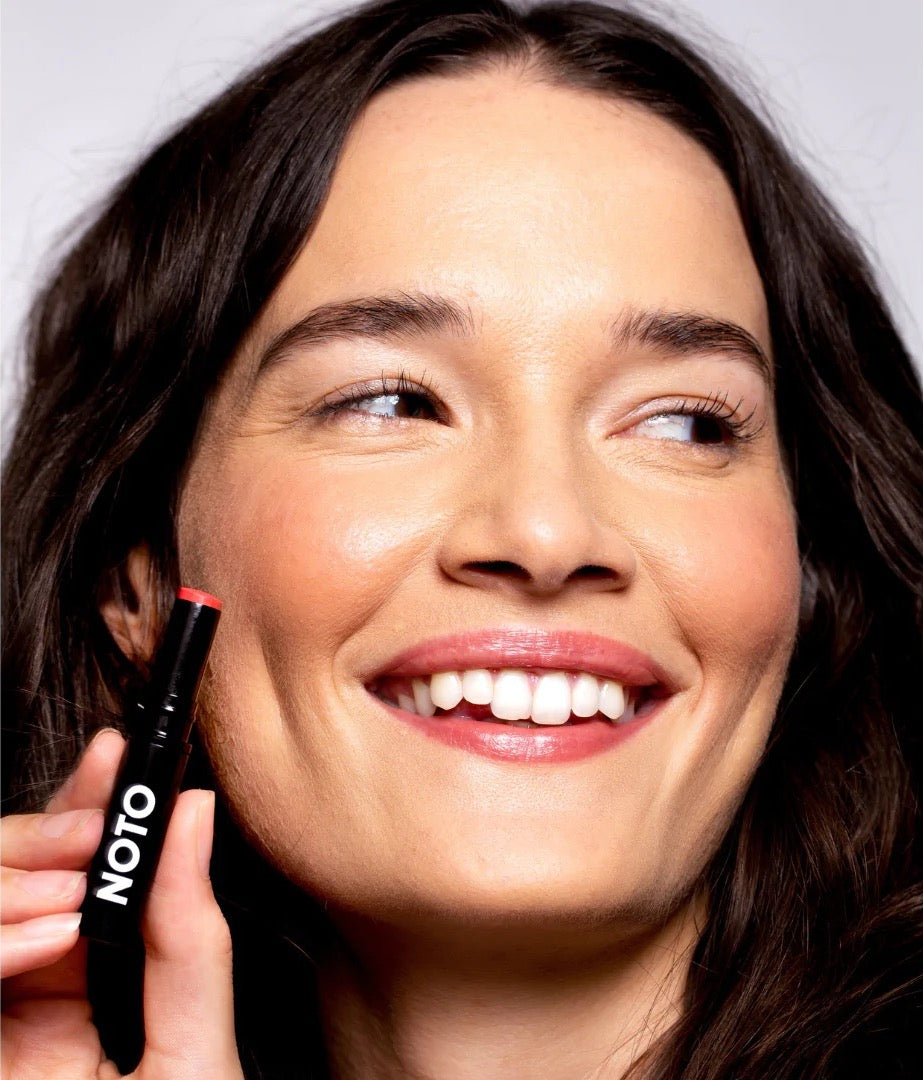 A woman is smiling while holding a NOTO Touch – Multi-Bene Stain Stick // Lips + Cheeks.