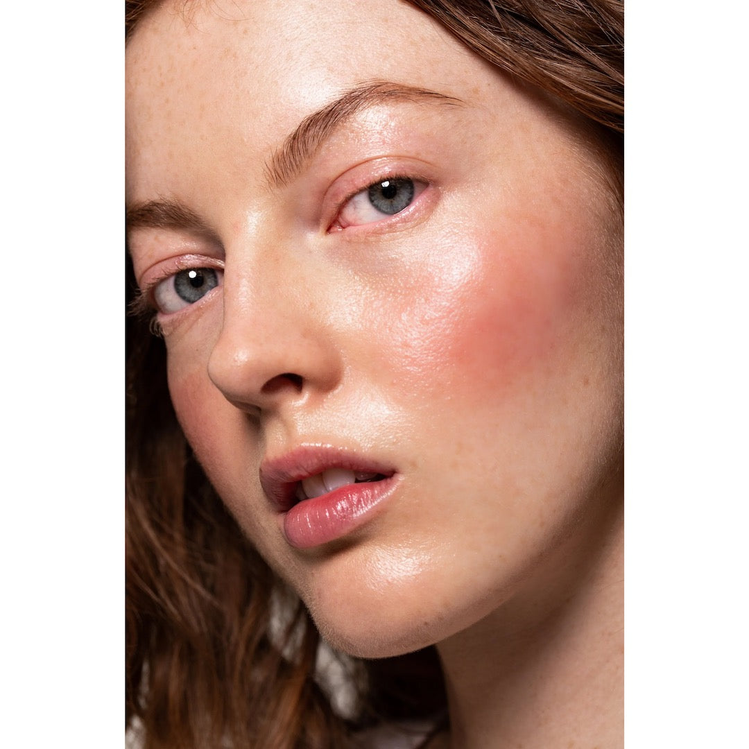 A woman with brown hair and a pink face using the NOTO Five – Multi-Bene Stain Stick // Lips + Cheeks.