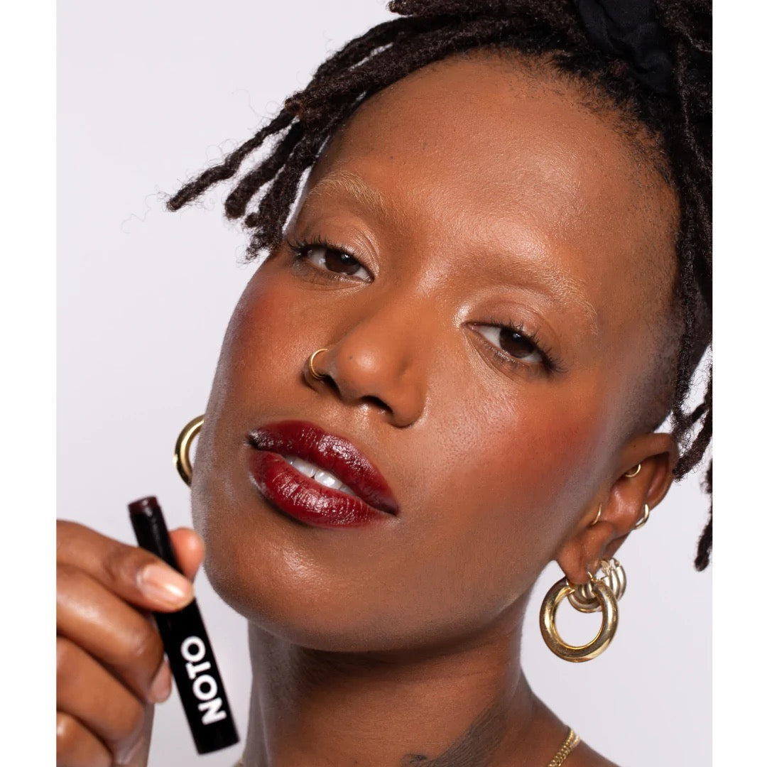 A woman with dreadlocks holding a NOTO – Genet Multi-Bene Stain Stick // Lips + Cheeks.