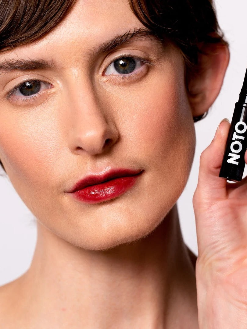 A woman is holding a Genet – Multi-Bene Stain Stick // Lips + Cheeks with a black tube, by the brand NOTO.