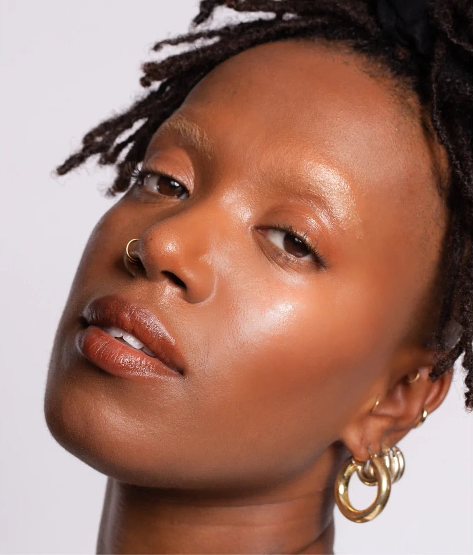 A black woman with dreadlocks and gold hoop earrings applied the NOTO Hydra Highlighter Stick // Face + Body.