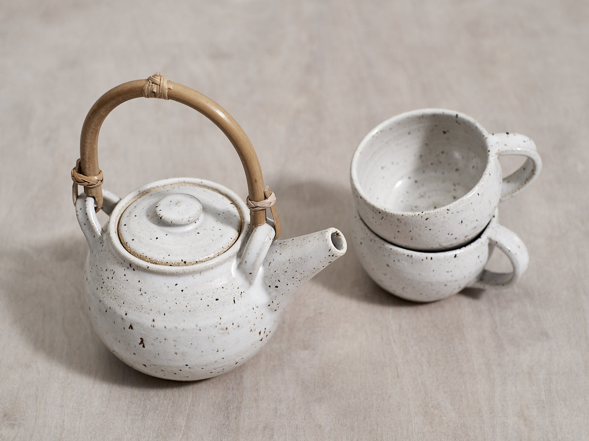 Two white handmade Nicola Shuttleworth speckled teapots and mugs with bamboo handles on a wooden table.