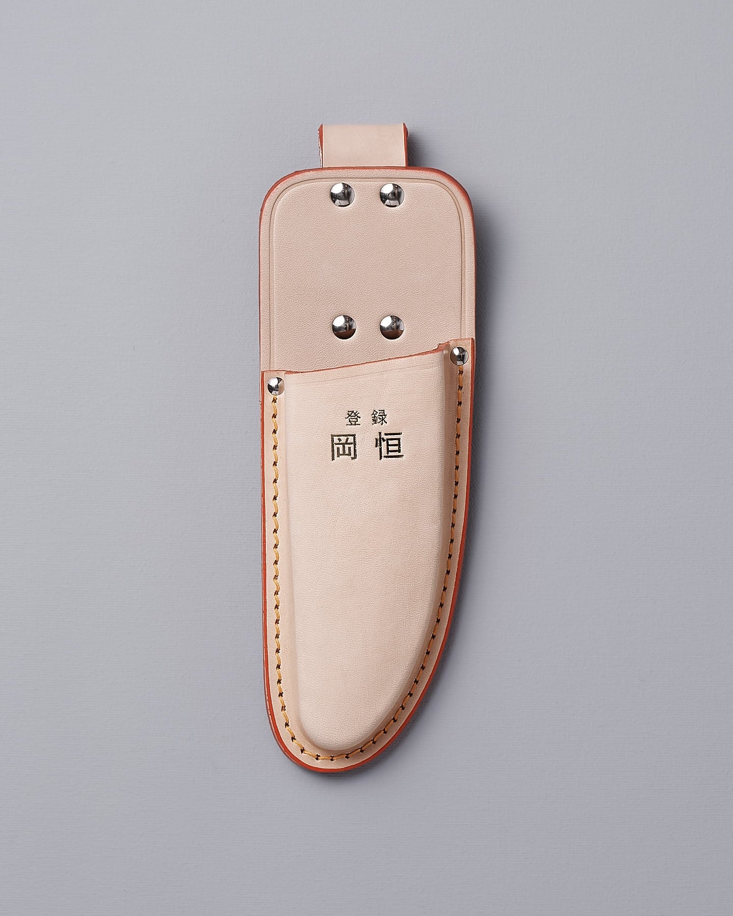 The Okatsune Japanese Secateurs №103 & Leather Holster – Bundle includes a beige leather holster adorned with metal rivets and a top loop, featuring Asian characters prominently on the front. It is expertly crafted to securely hold Japanese steel knives, all showcased against a gray backdrop.