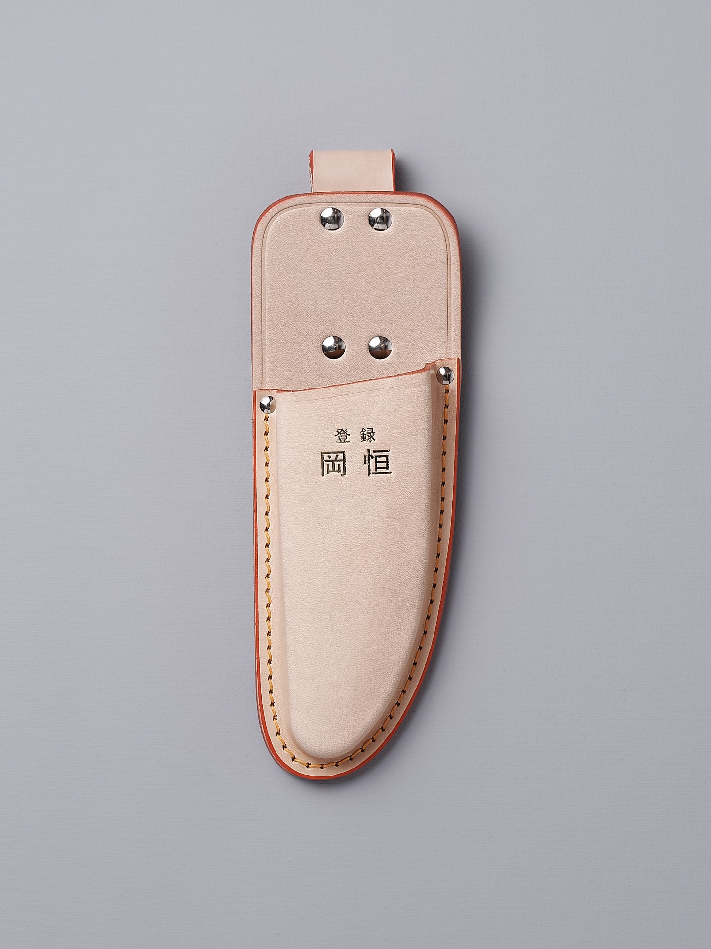 The Okatsune Japanese Secateurs №103 & Leather Holster – Bundle includes a beige leather holster adorned with metal rivets and a top loop, featuring Asian characters prominently on the front. It is expertly crafted to securely hold Japanese steel knives, all showcased against a gray backdrop.