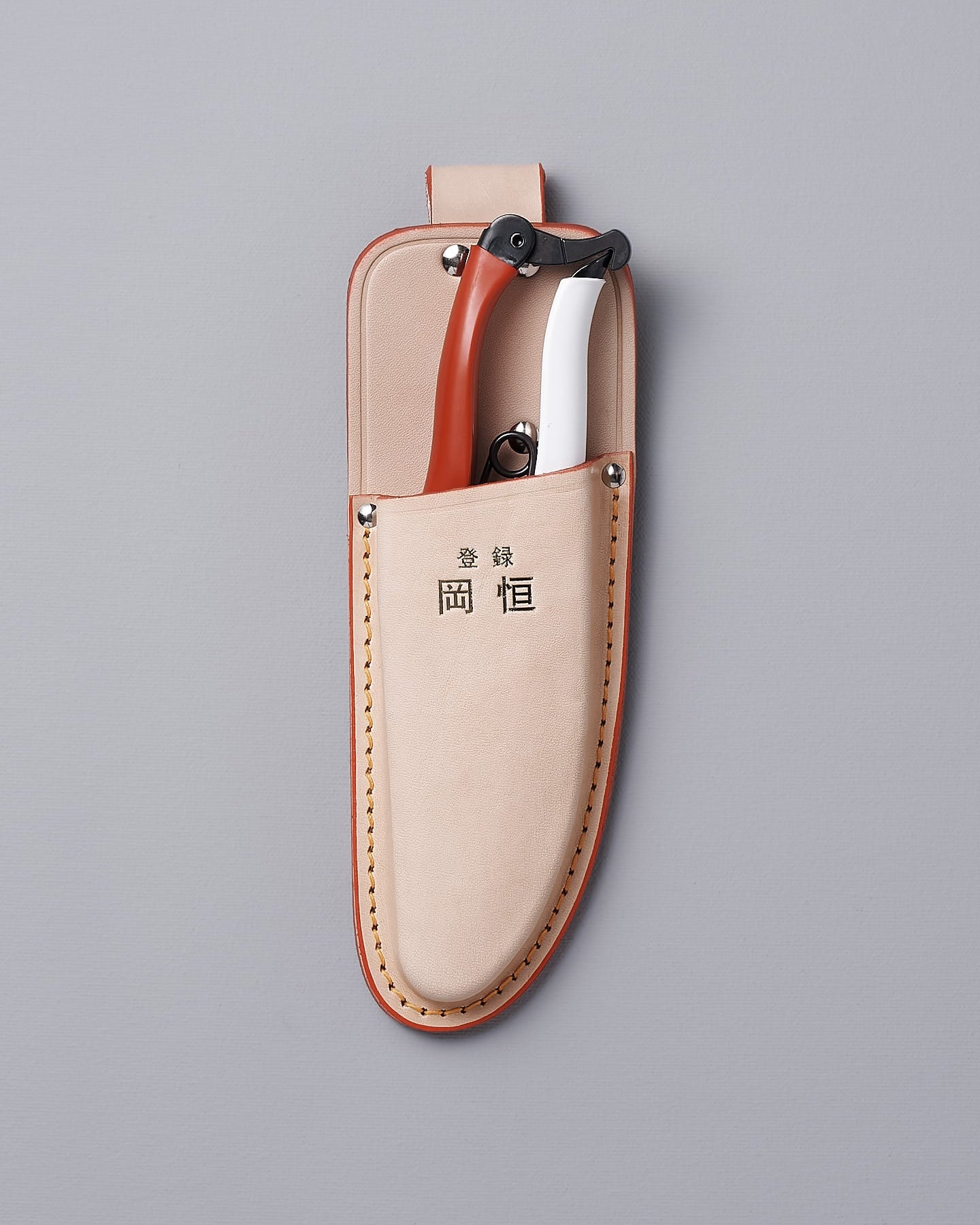 The Okatsune Japanese Secateurs №103 & Leather Holster – Bundle includes a beige leather holster adorned with metal rivets and a top loop, featuring Asian characters prominently on the front. It is expertly crafted to securely hold Japanese steel knives, all showcased against a gray backdrop.