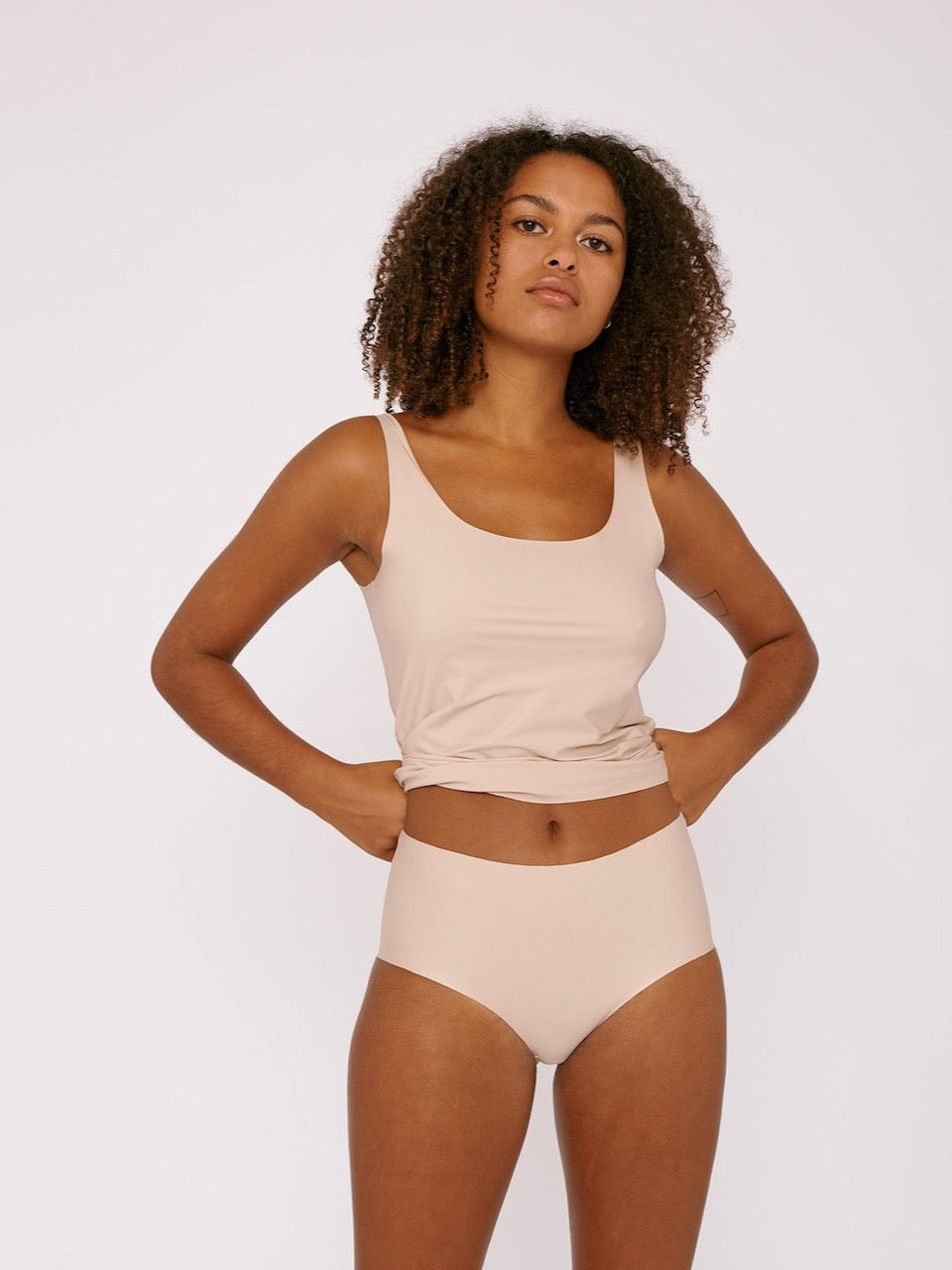 A woman in Organic Basics&#39; Invisible Cheeky High-Rise Briefs (2-pack) - Rose Nude tank top and panties.
