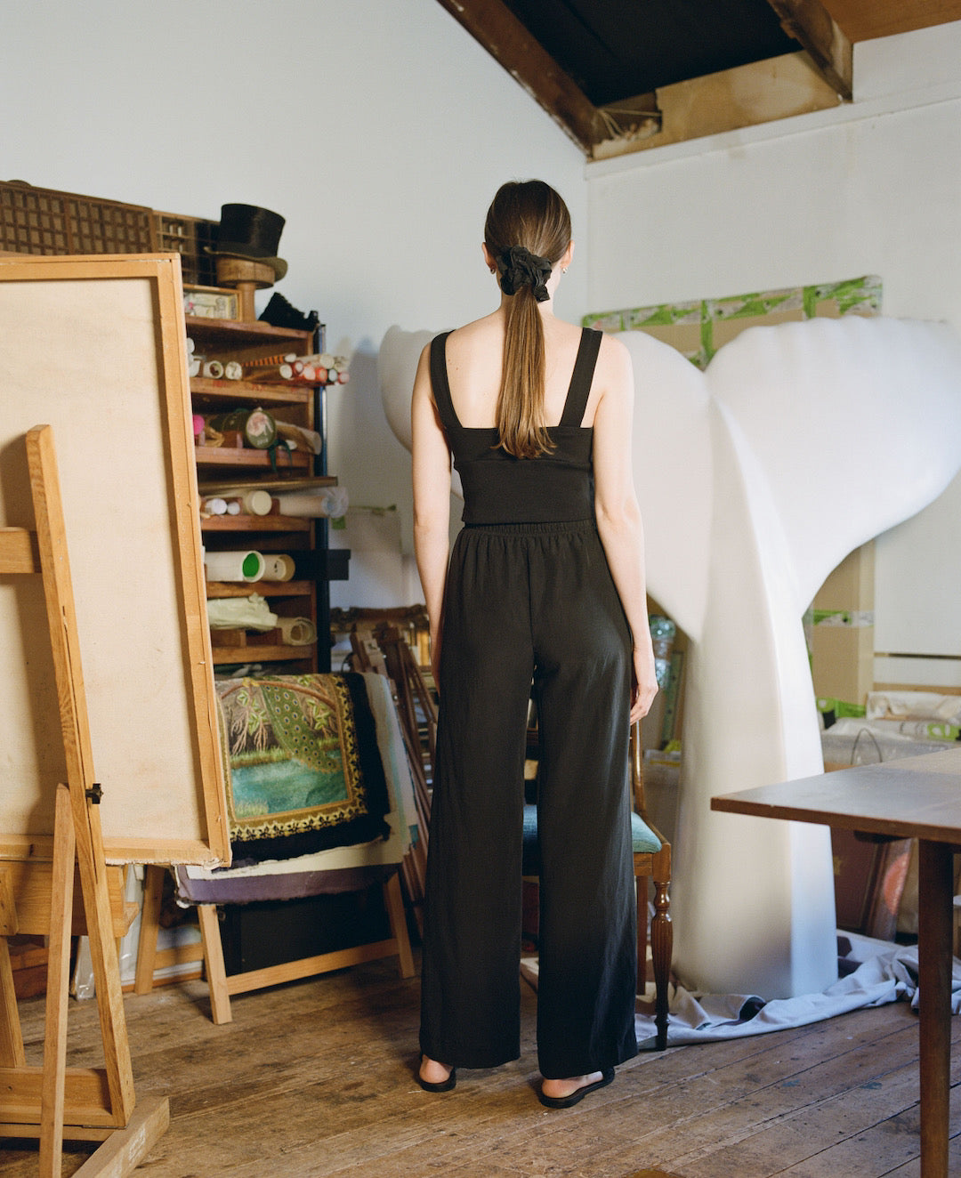 A woman in a black jumpsuit standing in front of an easel wearing the Eleanor Pant – Onyx by OVNA OVICH.