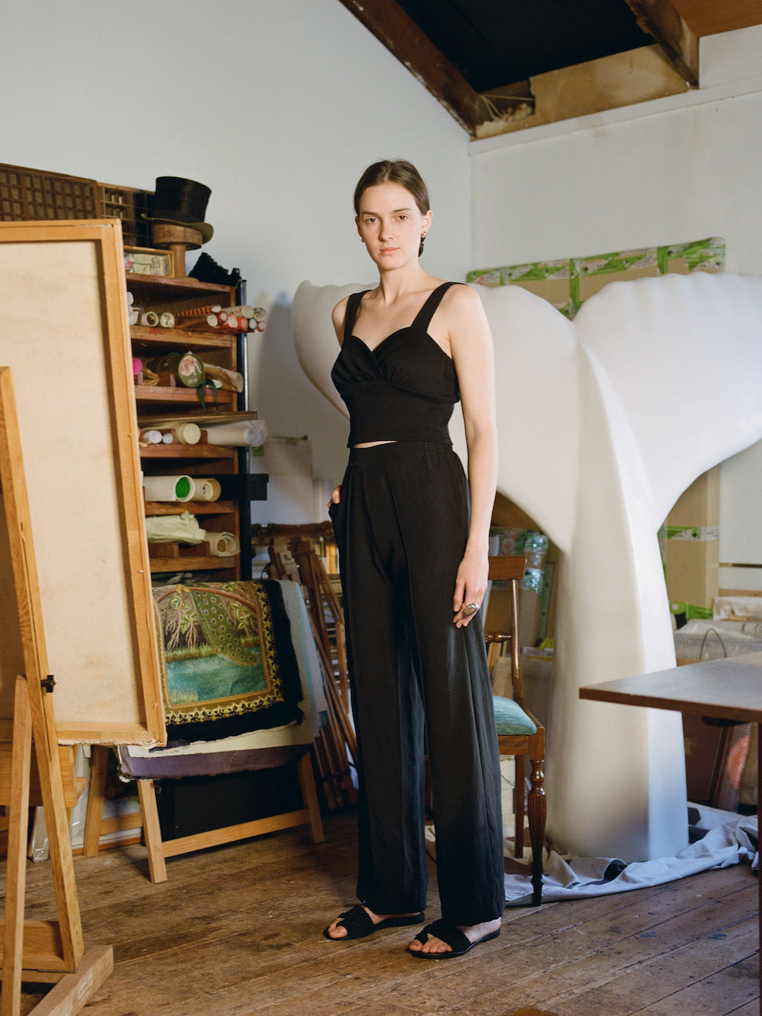 A woman in a black jumpsuit standing in front of an easel wearing the Eleanor Pant - Onyx by OVNA OVICH.