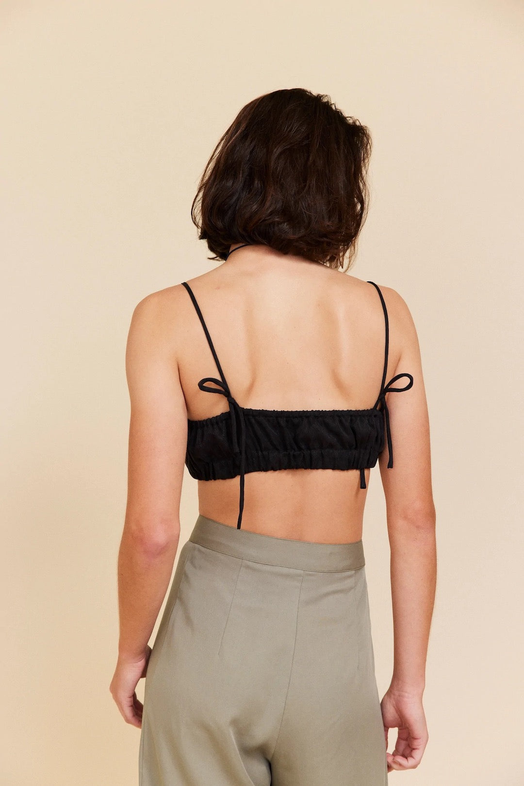 The back view of a woman wearing a OVNA OVICH Little Joy Bralette – Onyx and khaki pants.