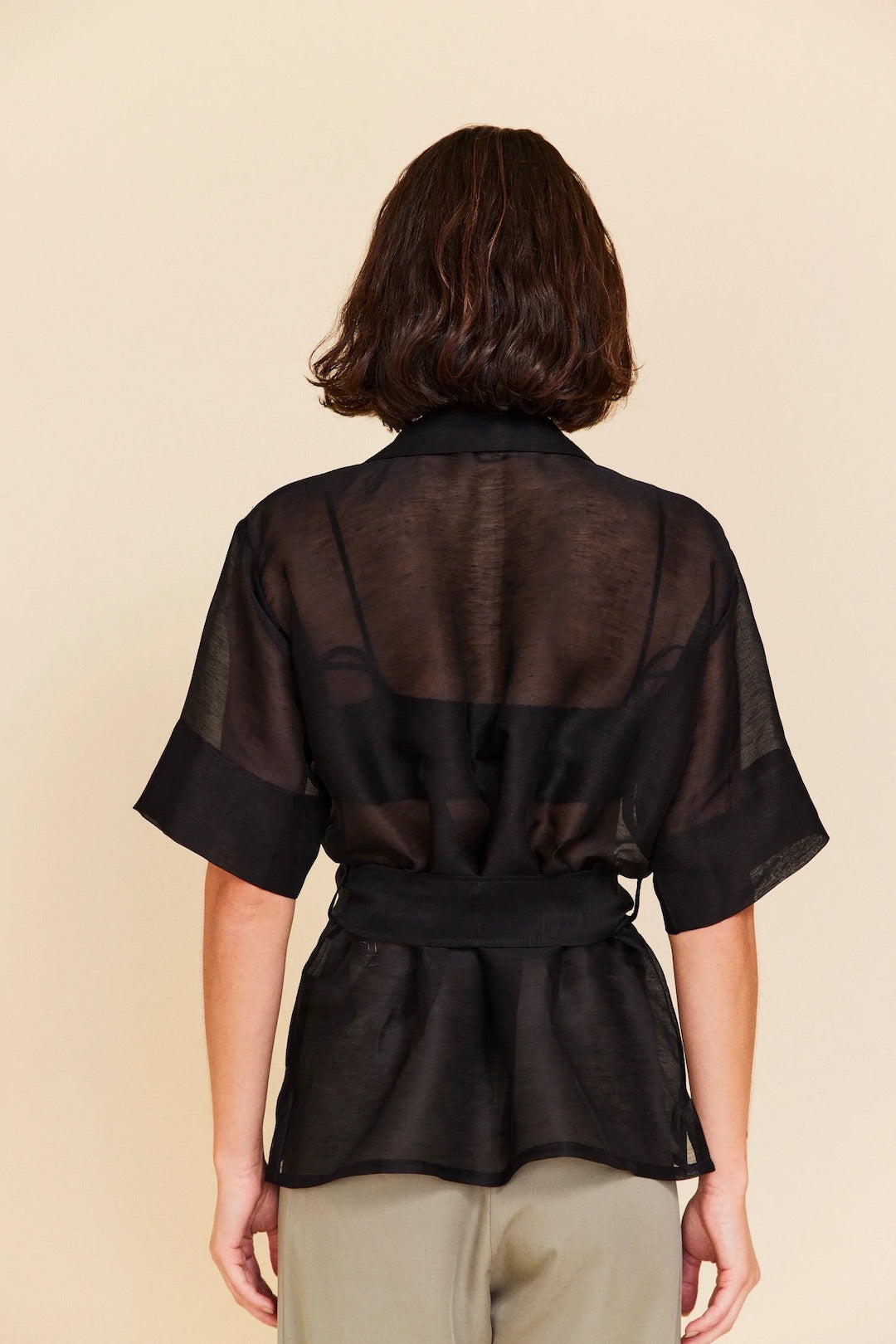 The back view of a woman wearing an OVNA OVICH Ora Shirt – Onyx and tan pants.