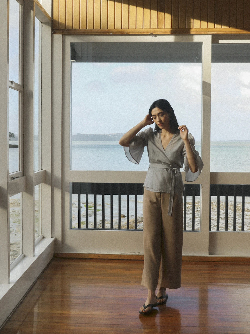 A woman standing in a room with a view of the ocean, wearing the Obira Wrap Blouse in Hazy Blue by OVNA OVICH.