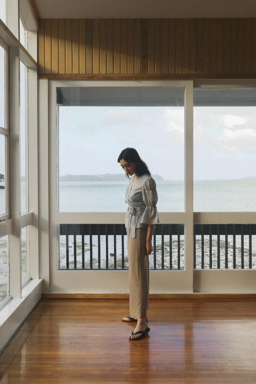 A woman standing in a room with a view of the ocean is wearing the OVNA OVICH Obira Wrap Blouse in Hazy Blue.