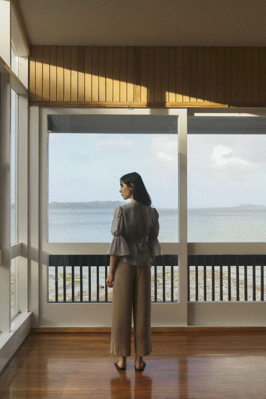 A woman standing in a room looking out to the ocean wearing an Obira Wrap Blouse in Hazy Blue by OVNA OVICH.