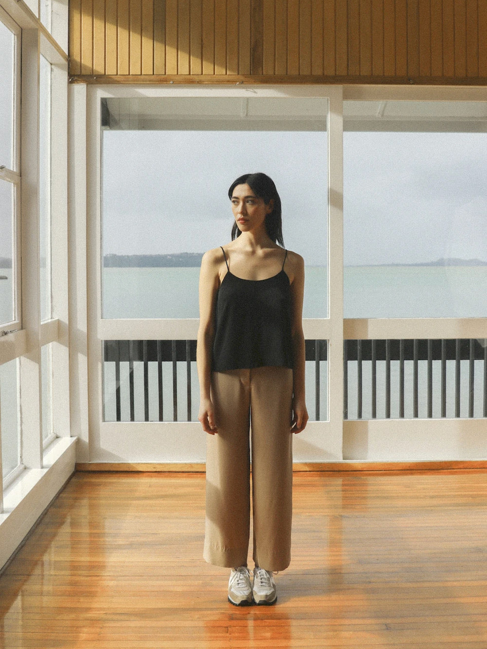 A woman standing in a room with a view of the ocean, wearing the Ya Pant - Taupe Tencel by OVNA OVICH.