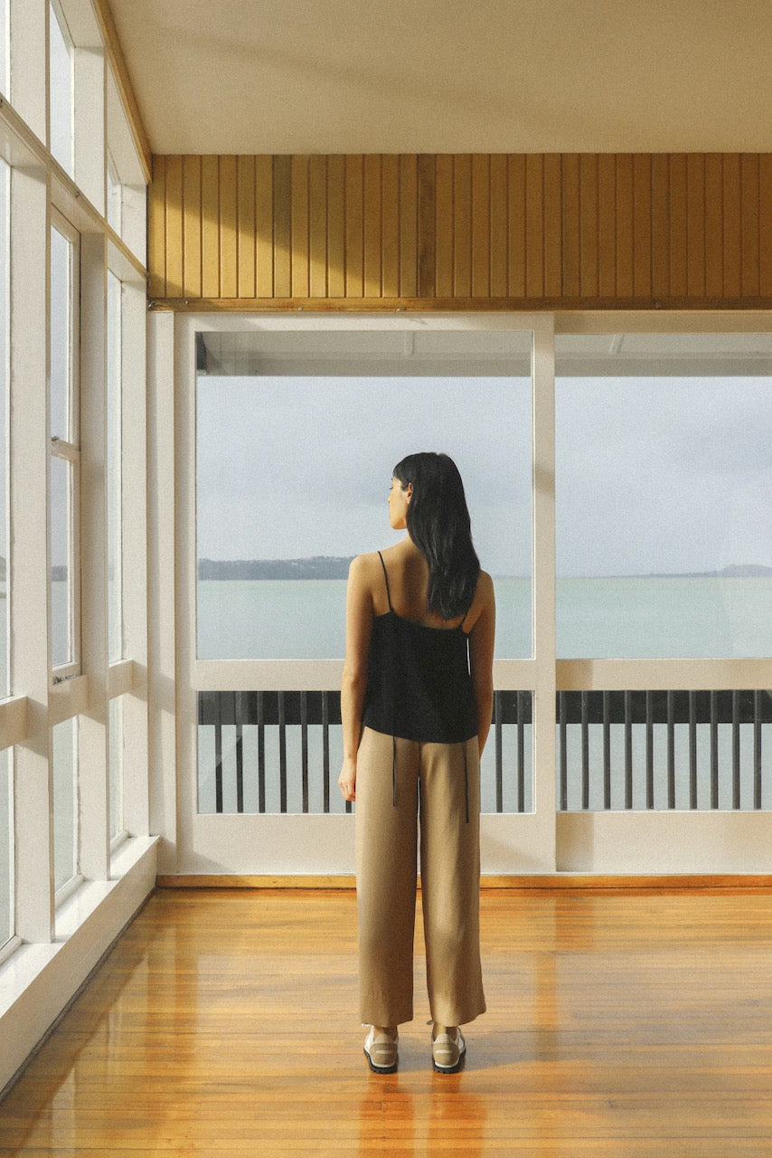 A woman standing in an empty room looking out to the ocean wearing the Ya Pant - Taupe Tencel by OVNA OVICH.
