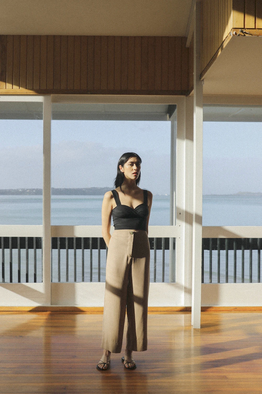 A woman standing on a wooden floor wearing the Ya Pant - Taupe Tencel by OVNA OVICH with a view of the ocean.