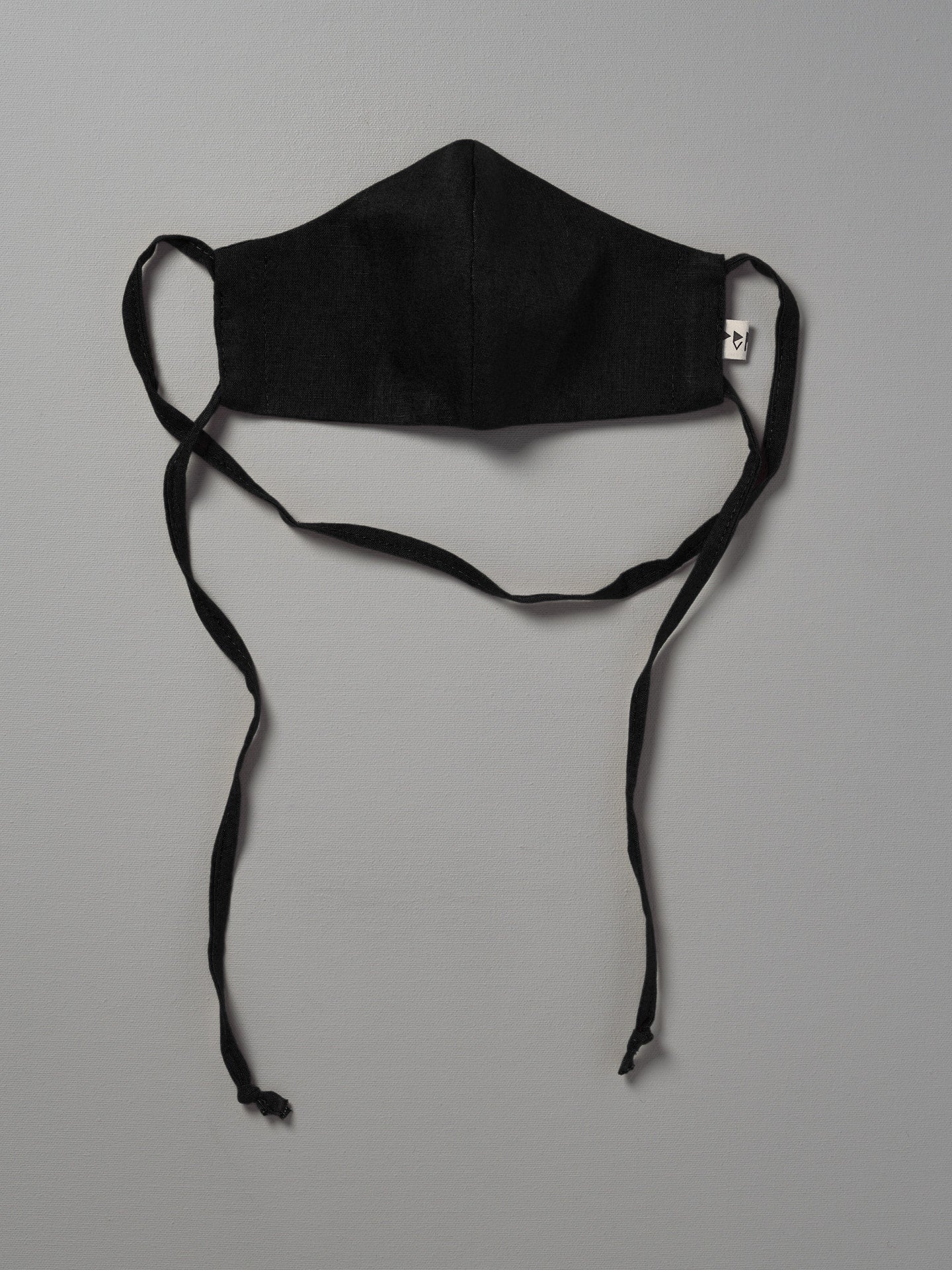A Linen Face Mask - Noir by Peppin hanging on a gray background.