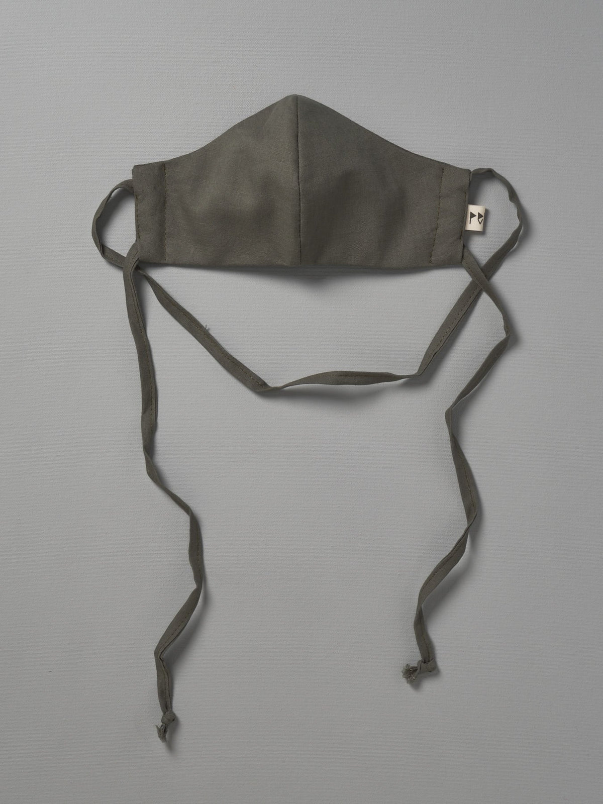 A Peppin Linen Face Mask - Moss hanging on a gray background.