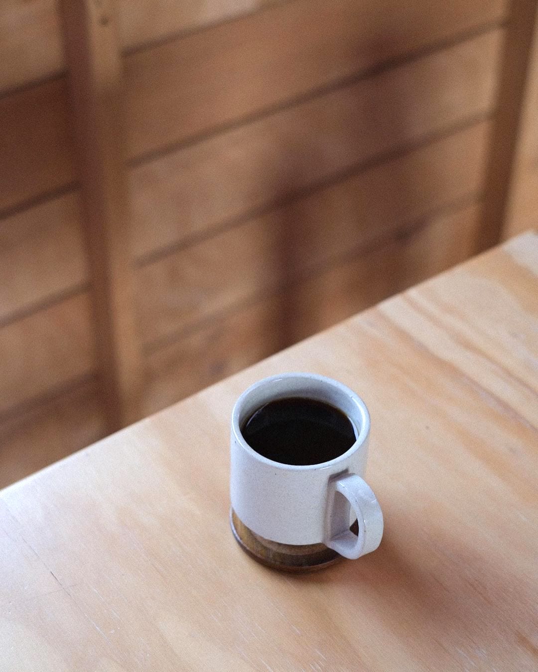 A Tall Stacking Mug – White by Richard Beauchamp, filled with black coffee, elegantly rests on a wooden table against a blurred wooden wall background in Ōtautahi Christchurch.