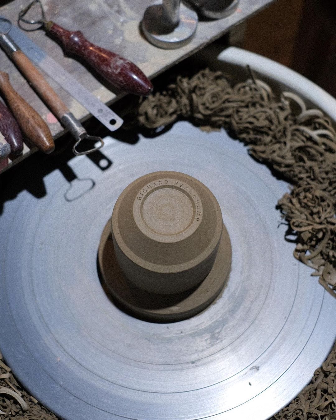 A Richard Beauchamp handmade potter's wheel on a table, accompanied by Richard Beauchamp ceramic stacking mugs (C-handled Stacking Mug – Sand) and various tools.