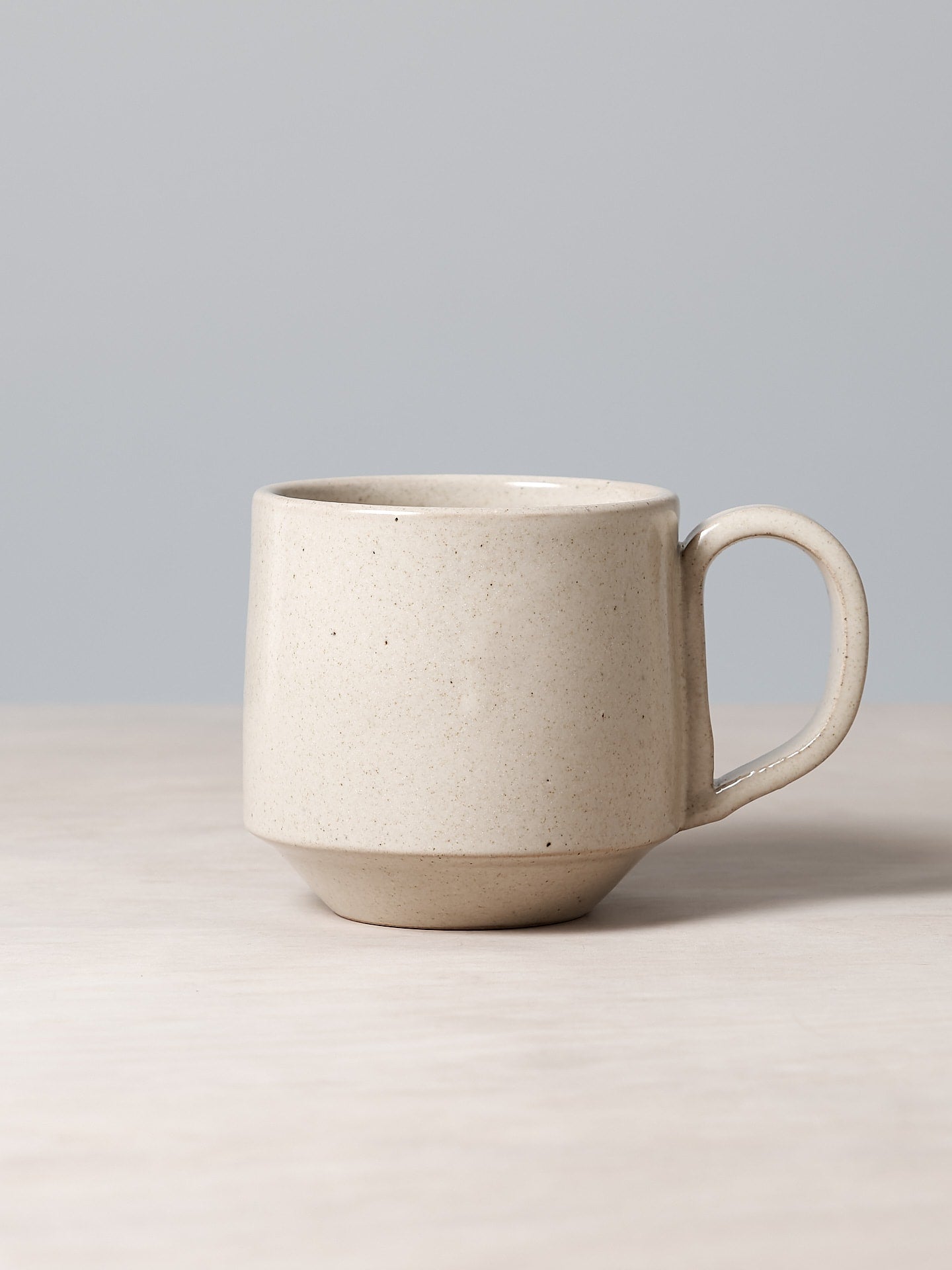Richard Beauchamp's Tall Stacking Mug – Beige, featuring a smooth finish, is gracefully displayed on a light wooden surface with a gray backdrop, highlighting its delicate beige glaze.