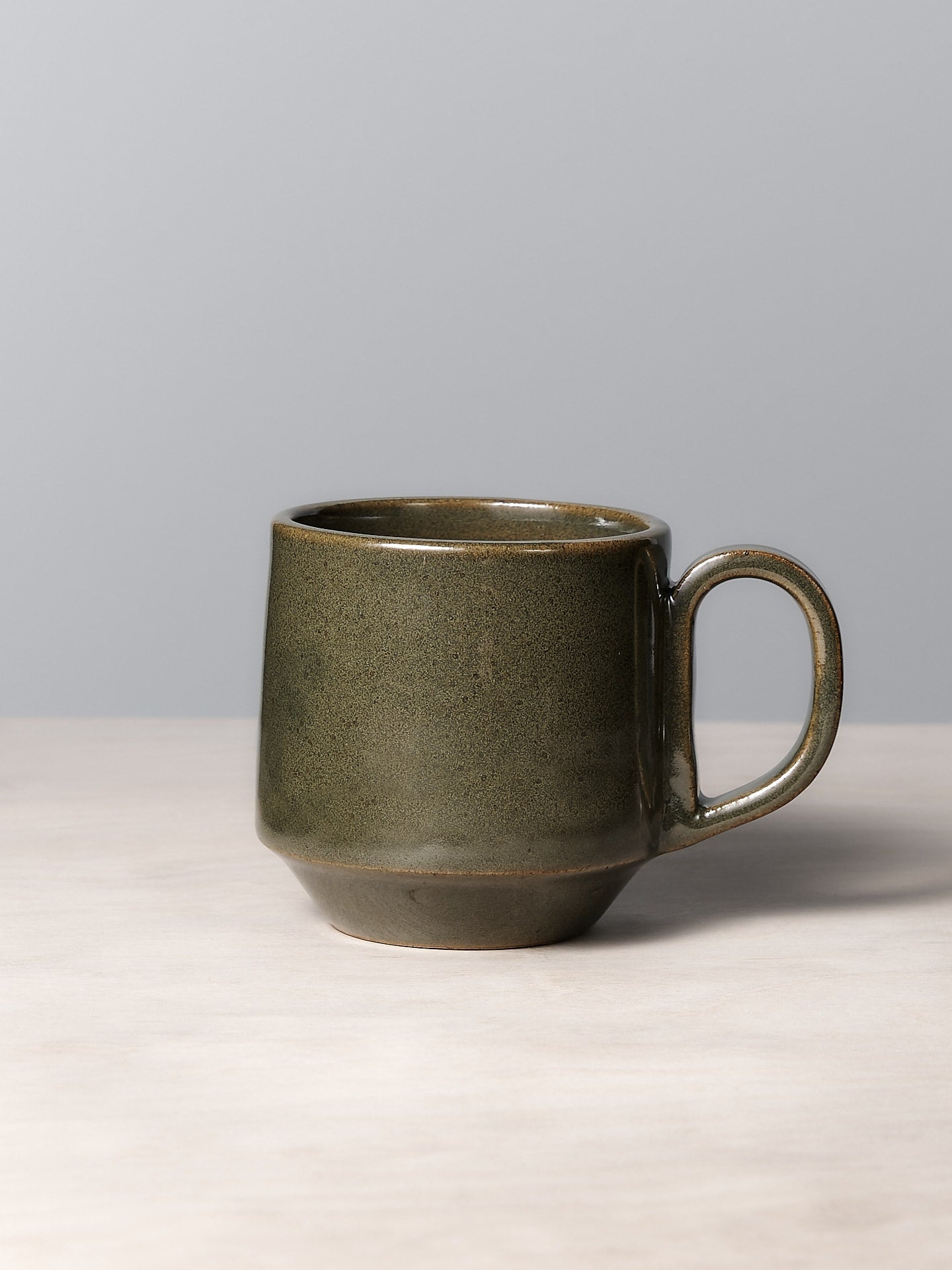 The Tall Stacking Mug – Green by Richard Beauchamp, boasting a smooth glaze finish, sits elegantly on a light wooden surface against a pale gray background.